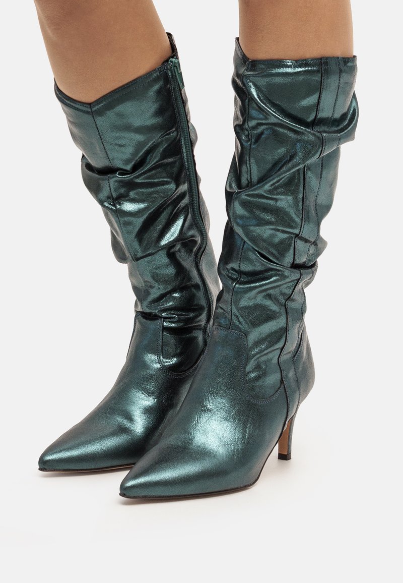 Bottes métalliques teal jusqu'aux genoux avec un bout pointu, des détails de fronces texturées et une fermeture éclair sur le côté ; elles possèdent un talon bas et fin.