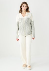 Veste zippée en polaire avec un corps texturé crème et gris, des manches crème et un col haut. Associée à un pantalon crème et des baskets beiges.