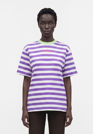 Kobieta ubrana w krótki rękaw, fioletowo-biały, poziomo paskowany T-shirt z zielonym kołnierzem i napisem „STIEGLITZ” na piersi.