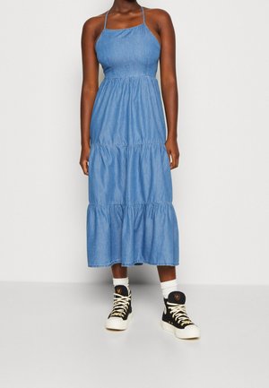 Denim-Maxikleid mit tailliertem Oberteil und gestuftem Rüschensaum. Hellblaue Farbe mit verstellbaren Trägern. Kombiniert mit schwarzen High-Top-Sneakers.