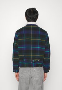 Chaqueta de cuadros en verde oscuro, azul marino y amarillo, con un cuello de borrego blanco y parte trasera ajustable con una tira. Tejido suave y corte entallado.