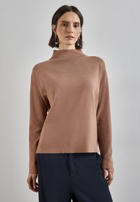 Kvinna klädd i en långärmad ljusbrun tröja med hög hals och mörkblå byxor, stående mot en enkel ljusgrå bakgrund.