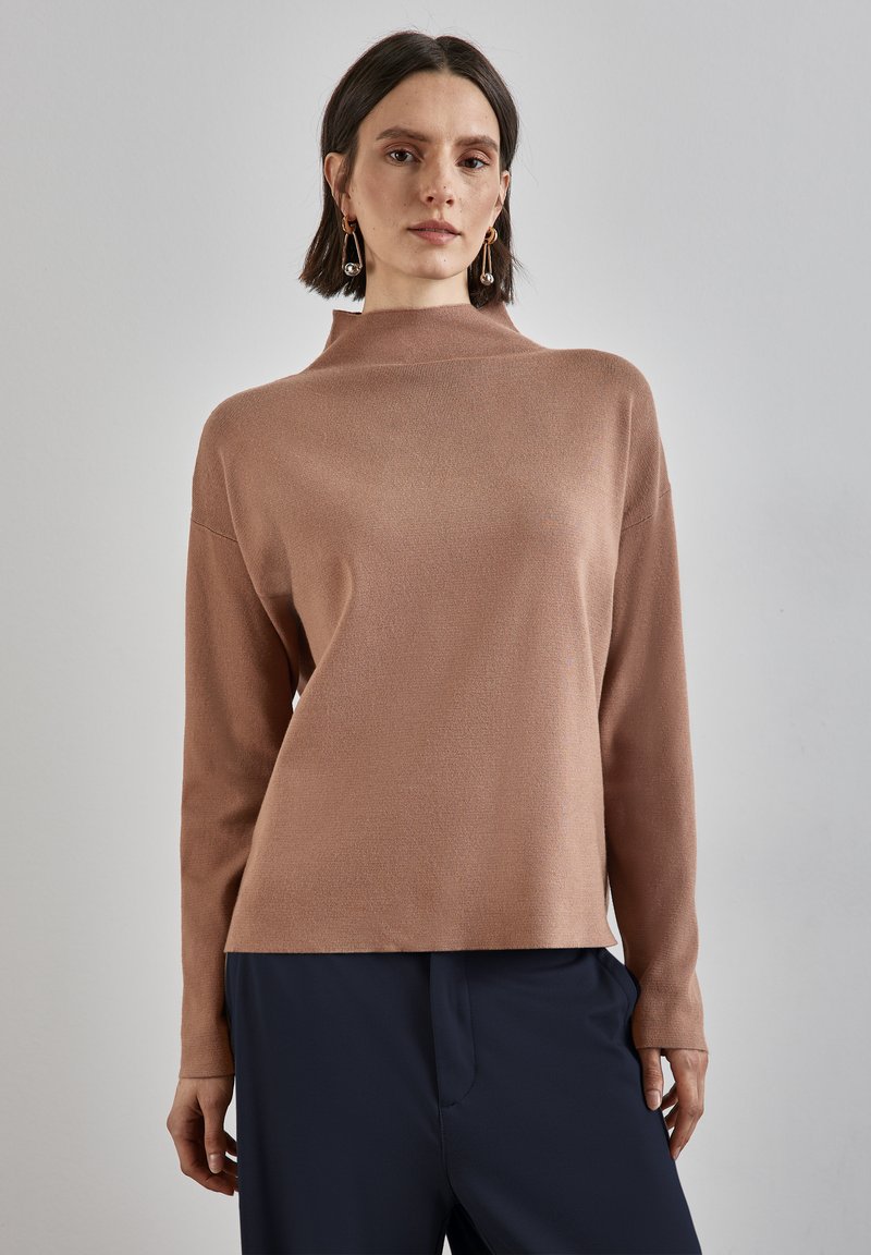 Kvinna klädd i en långärmad ljusbrun tröja med hög hals och mörkblå byxor, stående mot en enkel ljusgrå bakgrund.