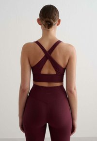 Burgundowy sportowy stanik z prążkowanej tkaniny z krzyżującymi się ramiączkami, zestawiony z legginsami o wysokim stanie. Gładka tekstura, minimalne logo na pasie.
