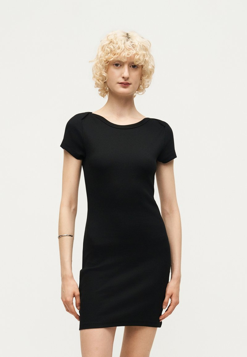 Γυναίκα με σγουρά ξανθά μαλλιά, φορώντας στενή μαύρη κοντομάνικη φόρεμα, στέκεται μπροστά σε ένα απλό φωτεινό φόντο.