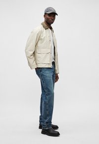 Δερμάτινο ελαφρύ μπουφάν σε μπεζ απόχρωση με καφέ γιακά, γκρι καπέλο, λευκή μπλούζα, μπλε τζιν παντελόνι και μαύρες μπότες. Παρουσίαση στυλ casual.