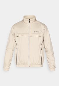 Bēša jaka ar augstu apkakli, priekšējo rāvējslēdzēju un melniem akcentiem. Uz krūškurvja ir logotips, un piedurknēm ir elastīgas aproces ciešai piegulšanai.