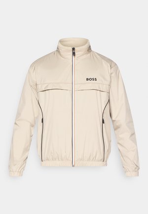 Μπεζ μπουφάν με ψηλό γιακά, μπροστινό φερμουάρ και μαύρες λεπτομέρειες. Διαθέτει λογότυπο στο στήθος και ελαστικά μανίκια για άνετη εφαρμογή.