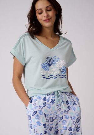 Donna con pigiama azzurro chiaro con disegno di conchiglie e pantaloni abbinati con motivi blu a tema mare, mani in tasca, occhi chiusi, leggero sorriso.