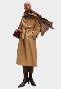 Lysebrun trenchcoat med bælte i dobbeltradet design, klaplommer og knapdetaljer; kombineret med et ternet tørklæde og en bordeaux taske.