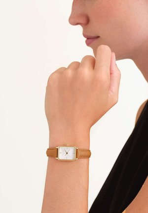 Montre carrée dorée avec un cadran blanc et un bracelet en cuir brun clair. Une main touche délicatement le cadran, mettant en valeur la montre au poignet.