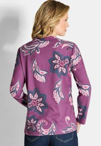 Frau mit schulterlangem blonden Haar, die ein lila Langarmoberteil mit großen rosa und marineblauen Blumen- und Blattmustern trägt, kombiniert mit dunkelblauen Jeans.
