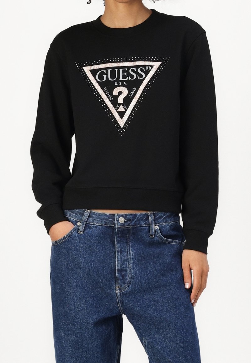 Personne portant un sweat-shirt noir avec un logo triangulaire Guess USA clouté en argent et un jean bleu, une main dans la poche.