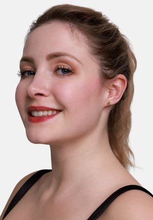 Goldene Steckerohrringe mit floralem Design. Das Thema hat helle Haut, trägt ein schwarzes Oberteil und hat die Haare zurückgebunden. Neutraler Hintergrund.