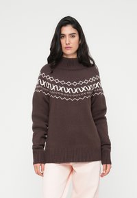 Καφέ πλεκτό πουλόβερ με ψηλό λαιμό και λευκό γεωμετρικό σχέδιο στο στήθος. Ριμπ μανσέτες και τελείωμα που δημιουργούν μια δομημένη εμφάνιση.