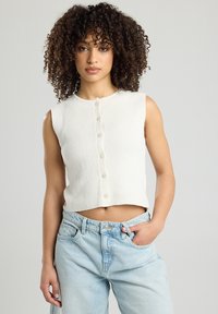 White ribbed sleeveless cropped top with a round neckline and six button closure down the front, paired with light blue high-waisted jeans.