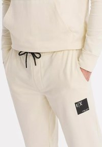 Cremfarbene Jogginghosen mit einem elastischen Bund, schwarzem Kordelzug, seitlichen Taschen und einem auffälligen schwarzen Etikett mit Logo.