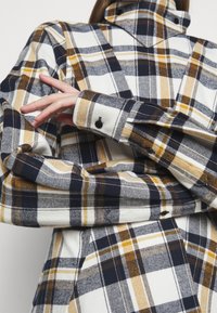 Plaid shirt in white, navy, and mustard. Features a high collar, long sleeves, and buttoned cuffs. Soft fabric with a textured finish.