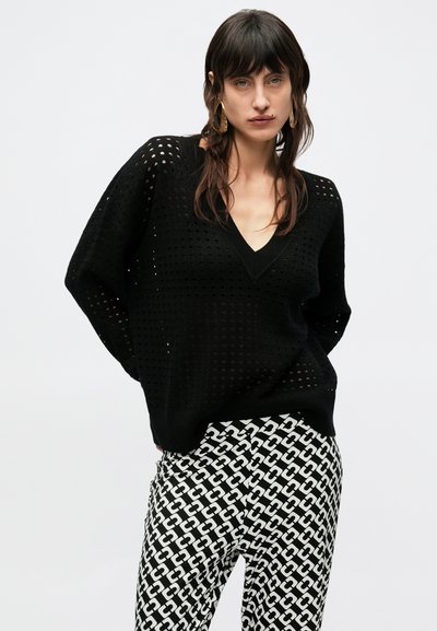 Must avara kudumiga kampsun V-kaelusega, kantud musta ja valge mustriga riiete kohal, millel on omavahel põimunud kujundid. Pikad varrukad, lõdva istuvusega.