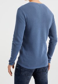 Långärmad blå tröja med en strukturerad, ribbad design. Har en rund halsringning och figursydda ärmslut. Bärs med blå jeans.
