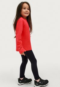 Helliges rotes Langarmoberteil mit rundem Ausschnitt, kombiniert mit schwarzen Leggings und schwarzen Turnschuhen mit weißen Sohlen, vor einem hellen Hintergrund.
