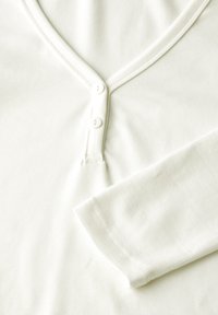 Ljust krämfärgad långärmad skjorta med V-hals, med två knappar vid kragen och ribbad struktur på tyget.