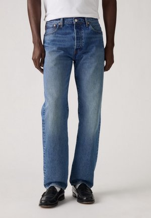 Mann, der blaue Jeans mit geradem Bein, ein weißes T-Shirt und schwarze Lederloafer trägt, steht vor einem schlichten hellen Hintergrund.