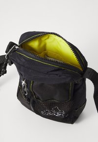 Černá taška s žlutým interiérem, se zipovým uzávěrem, síťovou kapsou a zeleným prošíváním. Obsahuje nastavitelný popruh pro pohodlné nošení.