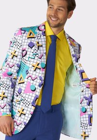 Blazer multicolore con fantasia a motivi, caratterizzato da una griglia bianca, accentuato da forme geometriche rosa, blu, gialle e viola. Camicia gialla brillante e cravatta blu.