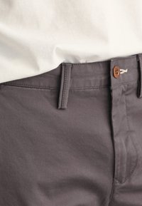Donkergrijze broek van een katoenmix, met een platte voorkant, achterzakken en een bruine knop bij de tailleband. Soepele textuur.