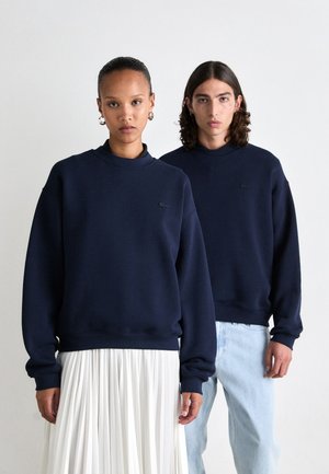 Navy blue cotton sweatshirts with ribbed cuffs and crew neck, featuring a small logo on the chest. Model wearing a white pleated skirt.