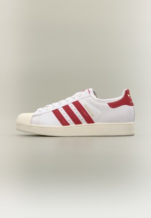 Witte Adidas Superstar sneaker met rode strepen, rode hieltab en gestructureerde neuskap op een neutrale achtergrond.