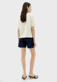 Witte top met korte mouwen gecombineerd met donkere denim shorts. Het model draagt open-toed zwarte sandalen met bontaccenten en een chunky zilveren ring.