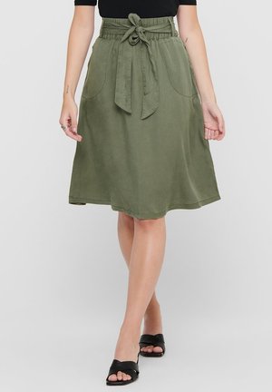 Femme portant une jupe mi-longue olive avec une ceinture nouée à la taille et des poches avant, associée à des sandales plates noires et un haut noir.