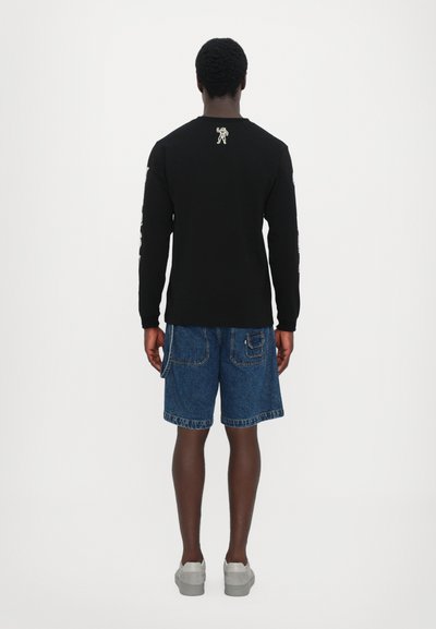 Sort langærmet skjorte med et lille hvidt grafik på ryggen, parret med blå denimshorts med sidelommer og en foldet kant.
