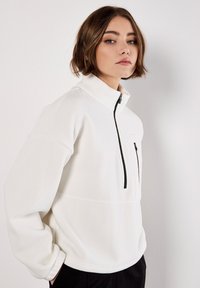 Hvid fleece-pullover med høj krave, halv-lynlås fortil og en brystlomme med sort lynlås. Blød tekstur og afslappet pasform.