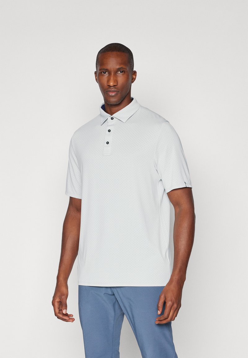 Hellgraues Poloshirt mit kurzen Ärmeln, strukturierter Musterung, drei Knöpfen und einem kontrastierenden Kragen. Kombiniert mit blauen strukturierten Hosen.