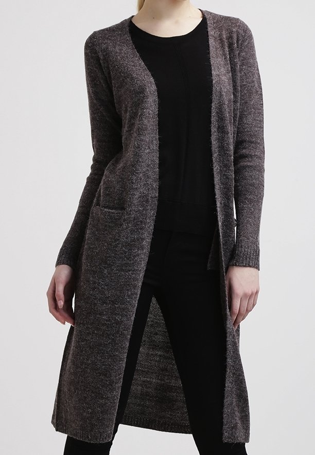 Femme portant un long cardigan gris foncé à ouverture frontale avec poches, sur un haut noir et un pantalon noir, se tenant devant un fond uni.