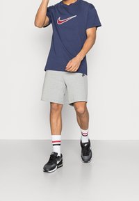 Námořnické bavlněné tričko s bílým a červeným logem Nike, doplněné o šedé kraťasy, černé tenisky a bílé ponožky s červenými pruhy.