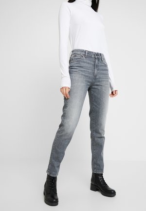 Person iført hvid langærmet rullekrave, grå højtaljede jeans og sorte snørestøvler ved anklerne, stående mod en ensfarvet baggrund.