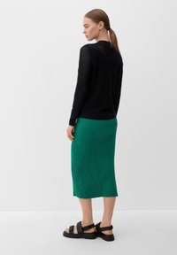 Zwarte opengehaakte trui met lange mouwen, gecombineerd met een groene geribbelde midi-rok. Zwarte chunky sandalen maken de outfit af.