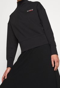 Sweat-shirt noir à col montant, détail brodé "LOVE" en blanc et rouge, avec manches longues et taille ajustée.
