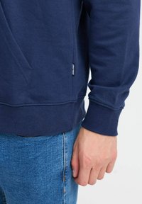 Navy blue cotton sweatshirt with a side pocket, ribbed cuffs, and waistband, featuring a small label near the hem. Paired with blue denim jeans.