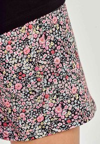 Krótkie spodenki w kwiaty mają czarne tło z gęstym wzorem różowych, żółtych i niebieskich kwiatów. Tkanina wydaje się miękka z delikatnym połyskiem.