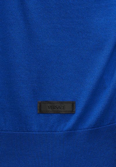 Μπλε πλεκτό ύφασμα με μια μικρή μαύρη ορθογώνια ετικέτα ραμμένη κοντά στο κάτω μέρος, με ανάγλυφο το λογότυπο «VERSACE».