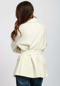 Giacca beige con cintura, dalla vestibilità morbida, design plissettato sul retro e tasche laterali. Realizzata in tessuto leggero e morbido.