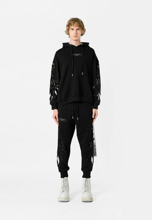Sudadera negra con detalles de flecos en las mangas, combinada con pantalones deportivos a juego. Cuenta con capucha, cordón y acentos negros en los bolsillos.