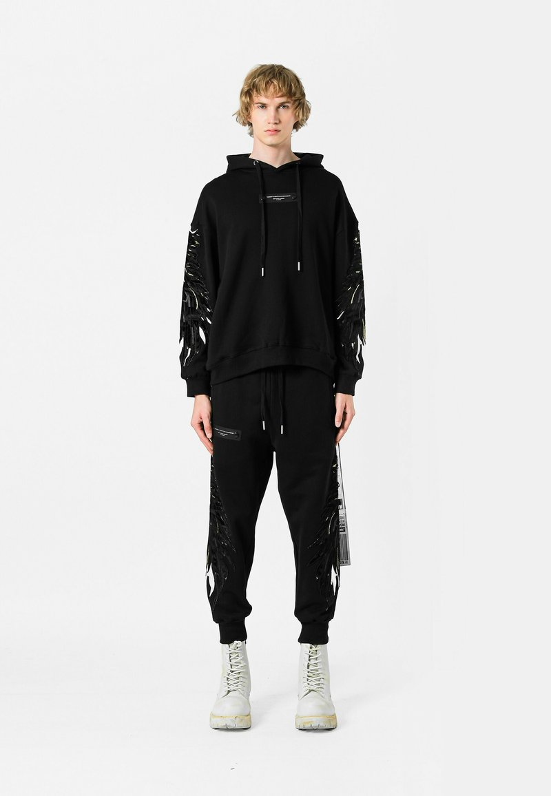 Sudadera negra con detalles de flecos en las mangas, combinada con pantalones deportivos a juego. Cuenta con capucha, cordón y acentos negros en los bolsillos.