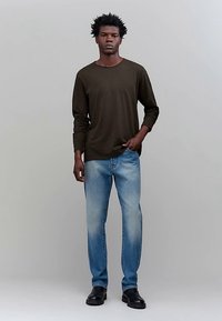 Dunkelgrünes Langarmshirt, hellblaue Jeans, schwarze Schuhe. Das Shirt hat einen runden Halsausschnitt; die Jeans sind leicht ausgewaschen. Das Modell steht gerade.