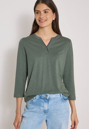 Frau mit langen dunklen Haaren, die ein grünes langärmeliges Shirt und hellblaue Jeans trägt, lächelnd vor einem schlichten Hintergrund stehend.